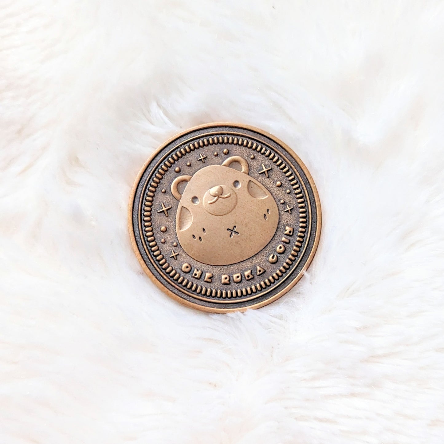 A photo of the front side of a coin made from zinc alloy with an antique gold finish. The coin features a bear in the middle and sparkle details around it. Under the bear it reads "One Ruka Coin".