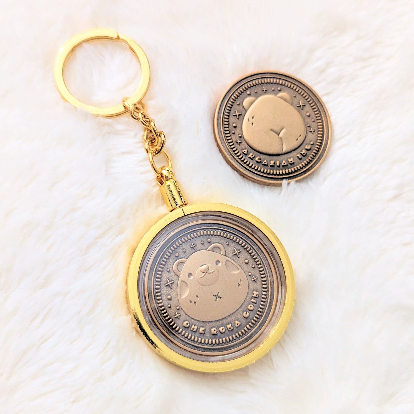 A photo of two coins made from zinc alloy with an antique gold finish. The coin of the left is inside a gold plated keychain attachment that shows the frontside of the coin with a bear design on it. The coin on the right is not enclosed in a keychain attachment and features the backside of the coin with the backside of the bear design showing.