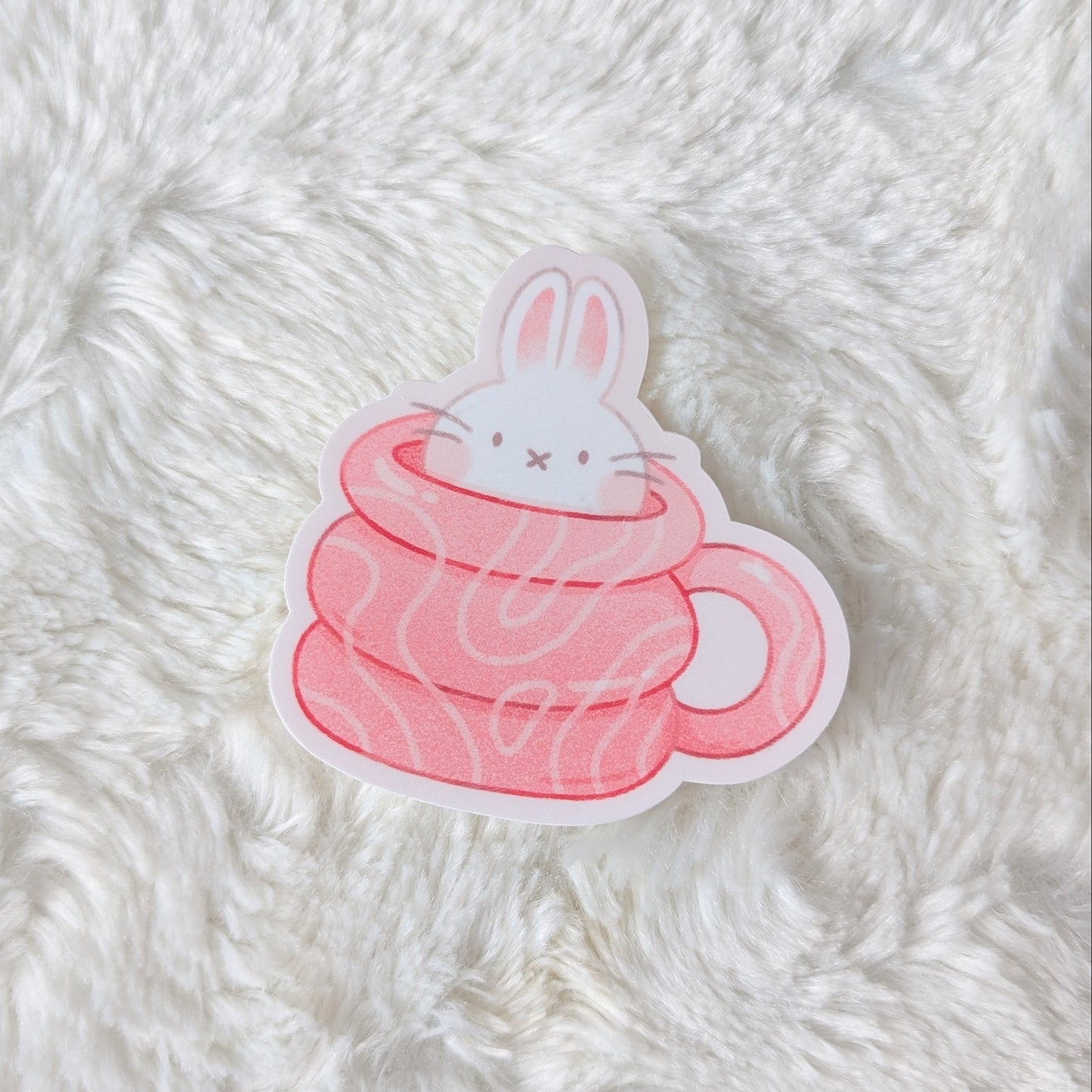 Sticker of a rabbit in a pink cup on a fluffy white surface
