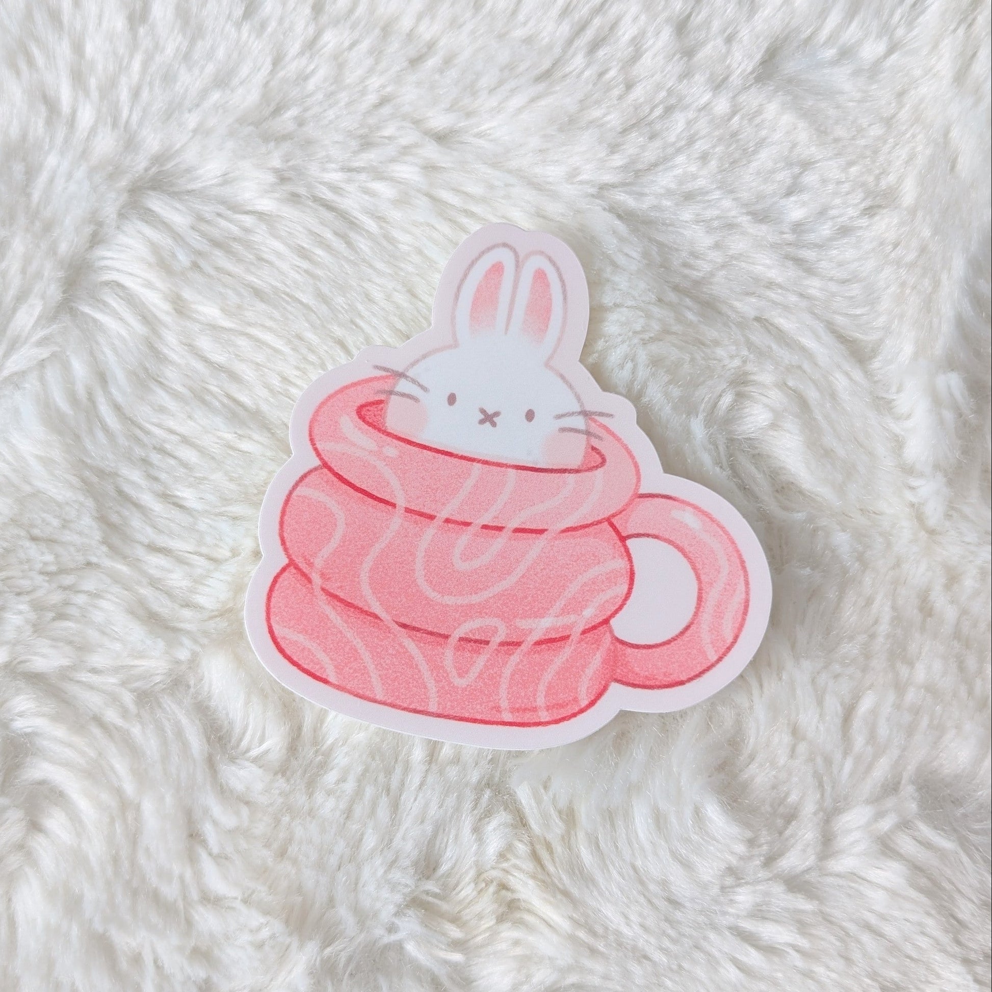 Sticker of a rabbit in a pink cup on a fluffy white surface