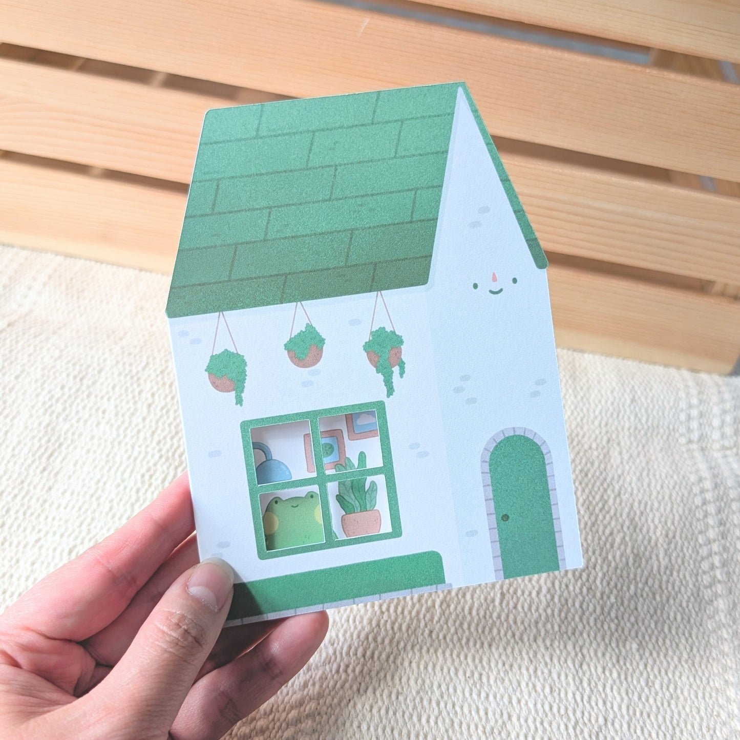 Hand holding a small green and white house-shaped card against a wooden background