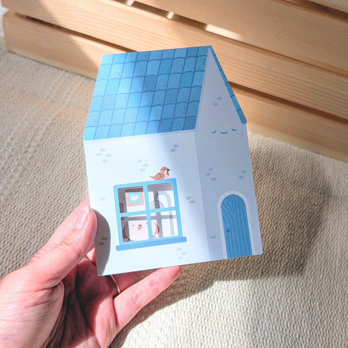 Hand holding a small house-shaped card with a blue roof and white body against a woven background.