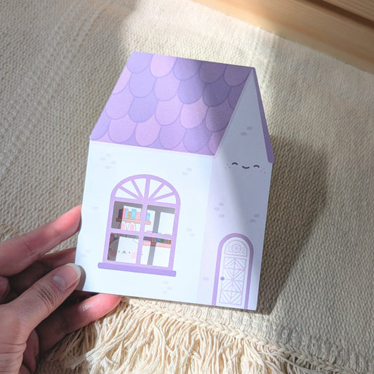 House-shaped card with purple roof and white body held by a hand on a textured surface.