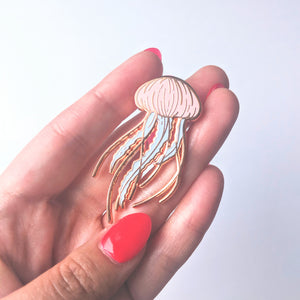 A close up of a rose gold jellyfish enamel pin being held by a hand with red nails
