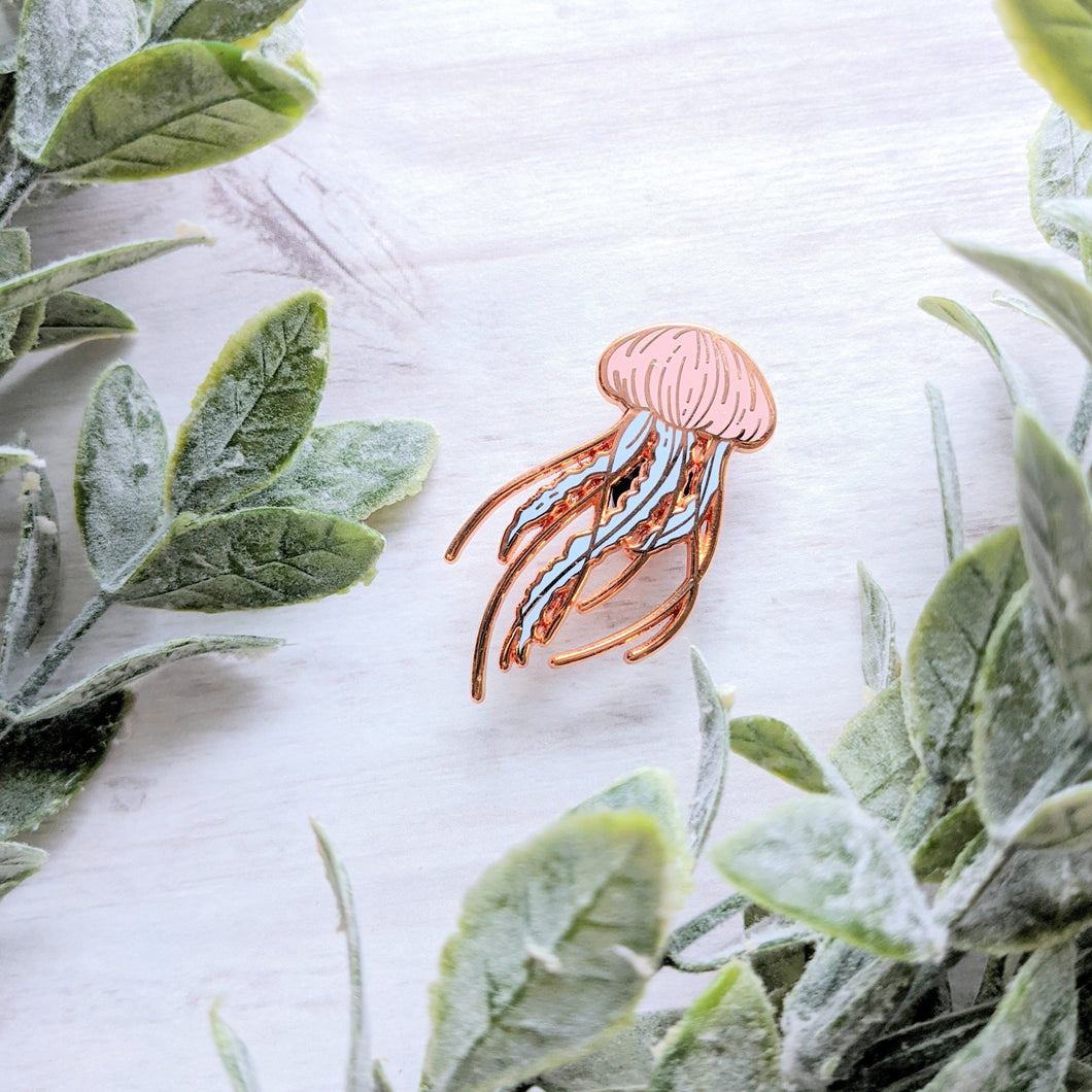 A rose gold pastel coloured enamel pin floating among small green leaves