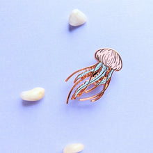 Load image into Gallery viewer, A single pastel rose gold jellyfish enamel pin amongst some small pebbles
