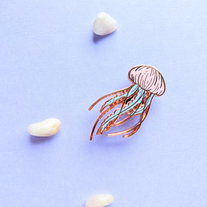 A single pastel rose gold jellyfish enamel pin amongst some small pebbles
