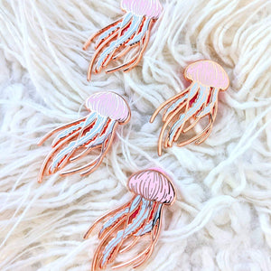 Four rose gold pastel jellyfish pins on a white textured pillow